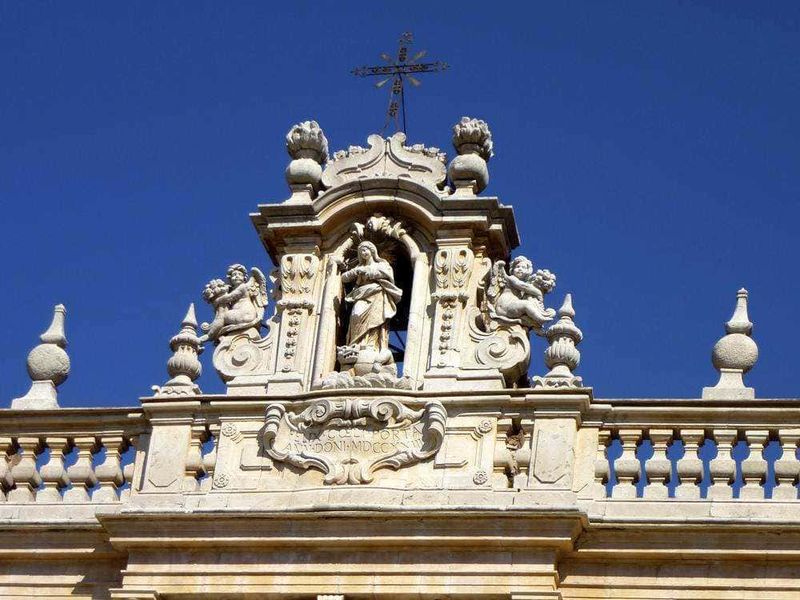 Ragazza scultorea in pietra decorativa sul tetto di un edificio storico, con un cielo azzurro sullo sfondo, elementi architettonici dettagliati e ornamenti barocchi, rappresentando arte e storia.