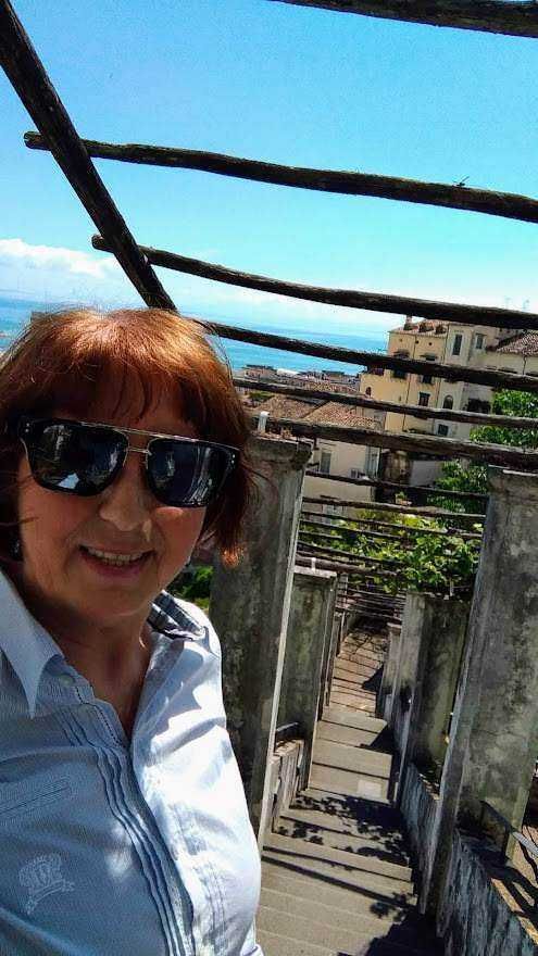 Appunti Sul Blog, donna sorridente con occhiali da sole sulla terrazza con vista panoramica, abbigliamento casual e sfondo di un cielo blu e edifici cittadini.