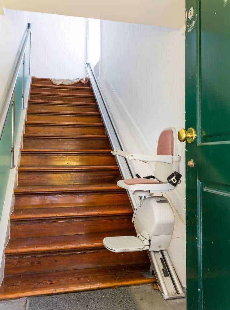 Scale di legno all'ingresso di un edificio con sedia mobile per disabili attaccata alla parete, vicino a porta verde. Spazio pubblico o aziendale con accesso facilitato per persone con mobilità ridotta.
