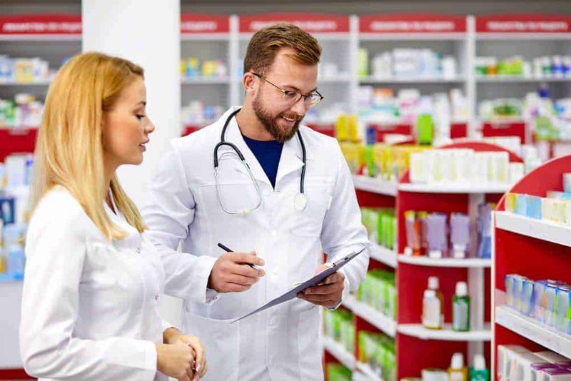L'importanza dei camici medici per un ambiente sicuro e pulito 3 Farmacisti in farmacia che consultano i farmaci sugli scaffali.
