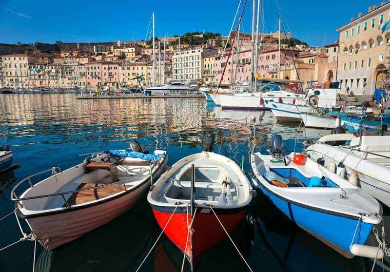 10 Attrazioni Isola d'Elba in 2 ore 3 Barche a vela attraccate nel porto di un pittoresco villaggio costiero, con edifici colorati sullo sfondo e acque calme riflettenti le imbarcazioni. Località di mare ideale per vacanze e nautica.