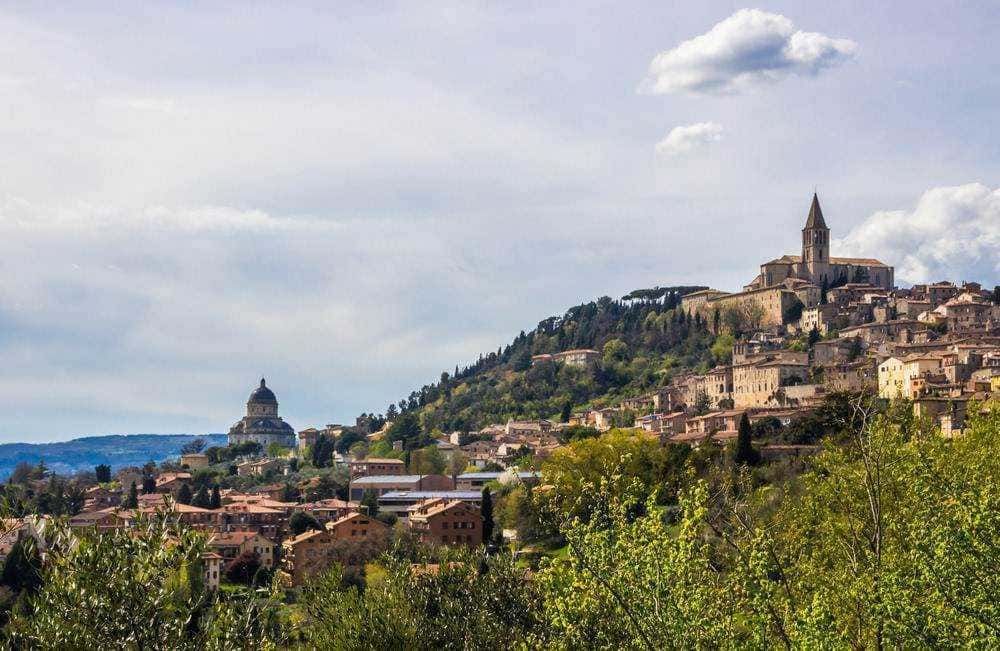 Vista panoramica di un antico borgo italiano su una collina con chiese storiche e case in pietra, circondata da natura e vegetazione verde, rappresenta un tipico paesaggio medievale in Italia.