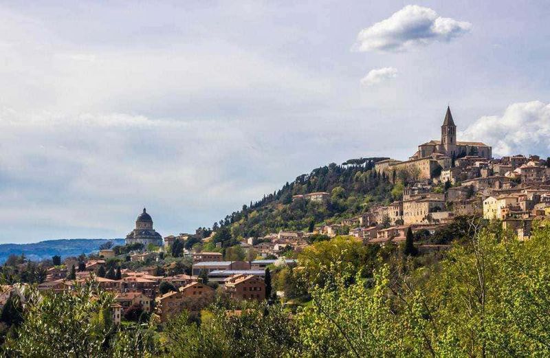 10 Città turistiche in Umbria da visitare, tra chiese romaniche 8 Vista panoramica di un antico borgo italiano su una collina con chiese storiche e case in pietra, circondata da natura e vegetazione verde, rappresenta un tipico paesaggio medievale in Italia.