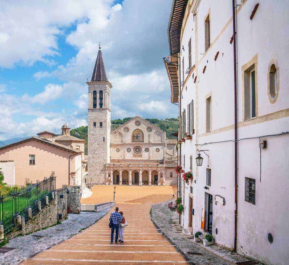 Chiese romaniche: Magnifico panorama di un borgo italiano con chiesa storica, cielo nuvoloso e coppia di turisti che passeggiano nel centro storico, perfetto esempio di turismo culturale e architettura tradizionale.