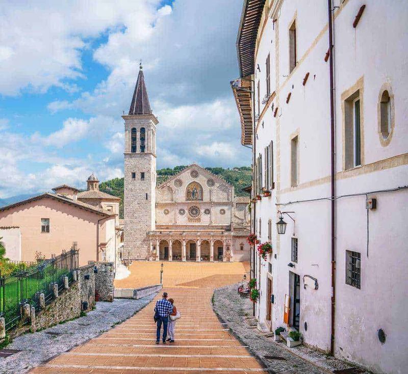 10 Città turistiche in Umbria da visitare, tra chiese romaniche 2 Chiese romaniche: Magnifico panorama di un borgo italiano con chiesa storica, cielo nuvoloso e coppia di turisti che passeggiano nel centro storico, perfetto esempio di turismo culturale e architettura tradizionale.