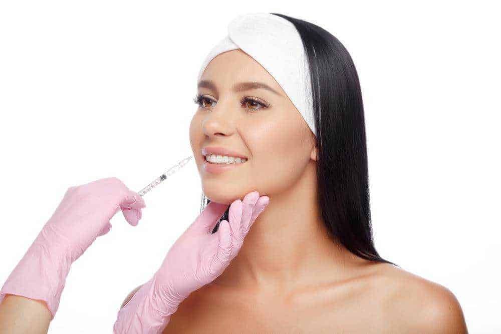 Belle donna che riceve un trattamento estetico con iniezione di filler o Botox, praticato da un'estetista con guanti rosa in un ambiente professionale, elegante e pulito.