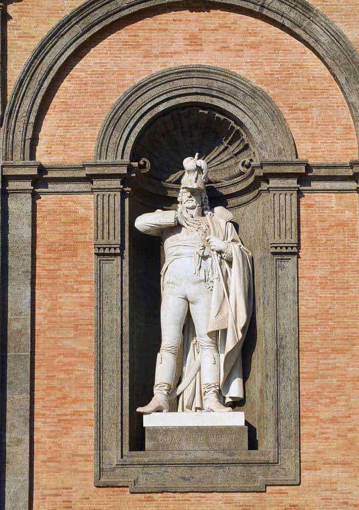 Statua in pietra di un uomo in abiti storici con un cappello ornamentale, parte di un monumento commemorativo su una parete di mattoni rossi, simbolo di cultura e storia italiana.