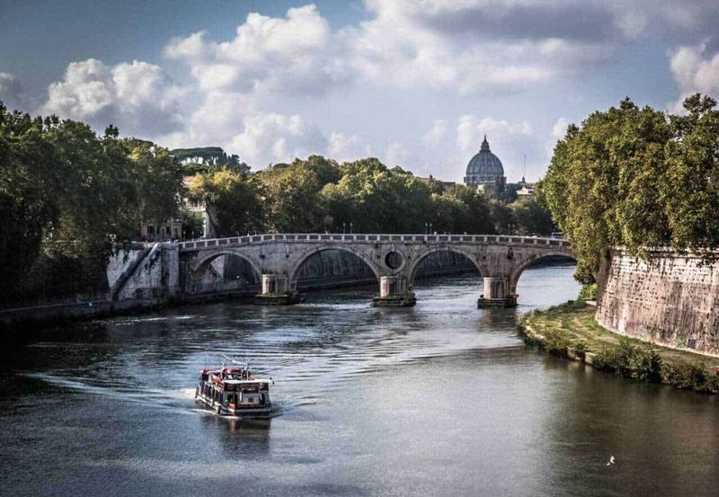 Roma in un lampo: deposito bagagli e come godersi la città in tempo record 2 Deposito bagagli: Pontes storiche sul Tevere a Roma con vista sul Vaticano e cielo nuvoloso. Nave che naviga sul fiume, circondato da alberi e edifici storici. Perfetta immagine di Roma e il suo patrimonio architettonico.