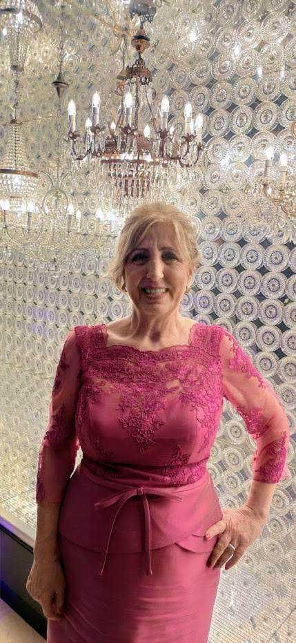 Elegante donna in abito rosa con decorazioni in pizzo, sorridente, in ambientazione di lusso con lampadari chandelier e pareti decorative con motivi geometrici, evento raffinato.