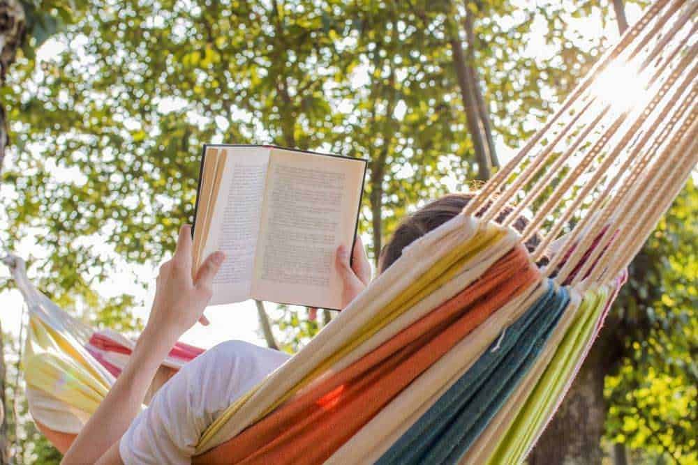 Relaxing in a hammock outdoors while reading a book on a sunny day, surrounded by green trees, embodying leisure and outdoor reading experiences.