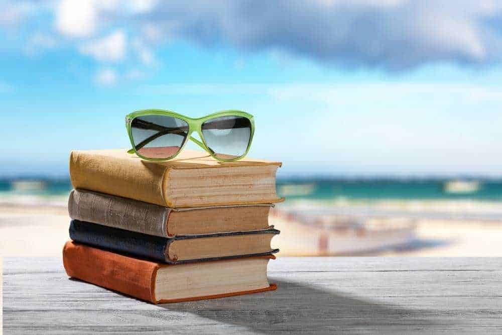 Vari sunglasses da spiaggia su un pilastro di libri, con sfondo mare e cielo azzurro, ideale per articoli sul relax, vacanze estive e lettura sotto il sole.