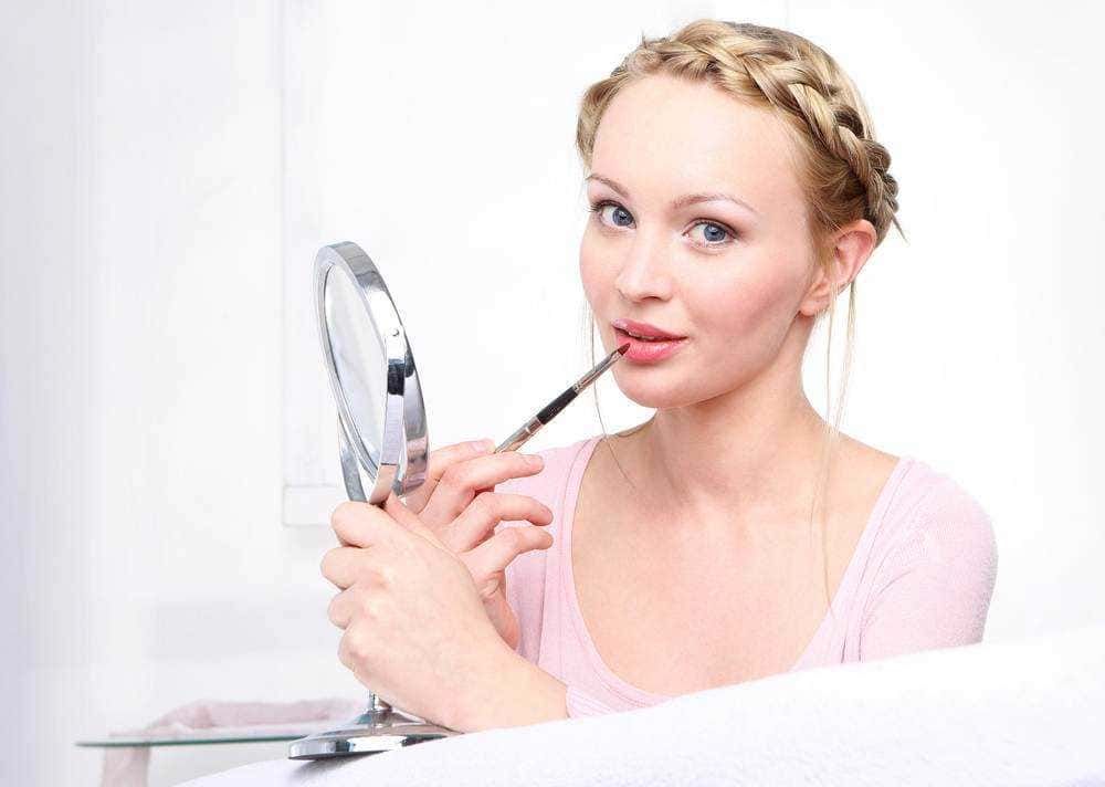 Bellissima donna che si trucca con il gloss, usando un pennello per applicarlo sulle labbra, con uno specchio da trucco davanti, in un ambiente luminoso e pulito, ideale per contenuti di bellezza e make-up.