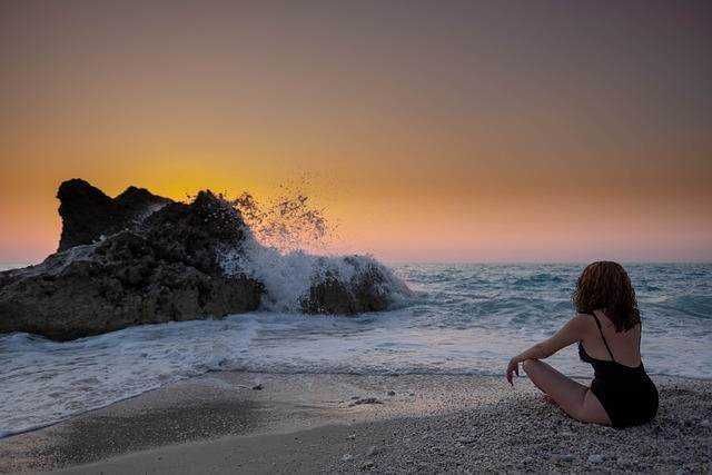 Tranquilla donna seduta sulla spiaggia al tramonto con onde che si infrangono su una roccia, atmosfera serena e relax al mare.
