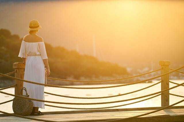 Vecchia donna con cappello e vestito bianco che guarda il tramonto dal pontile sul fiume in un paesaggio naturale, atmosfera serena e rilassante.