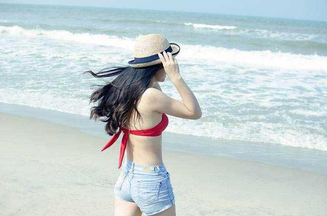 Vacanze al mare con stile: Donna in costume da spiaggia che guarda il mare sulla spiaggia, indossando cappello di paglia, costume rosso e shorts in denim, atmosfera estiva e relax.