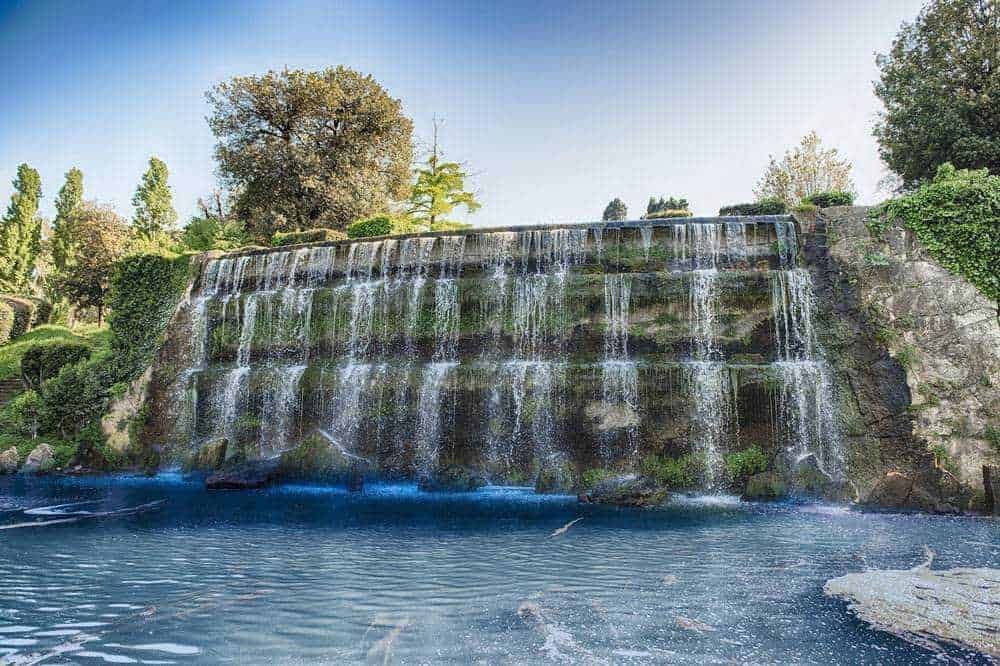 Serpente d'acqua che scorre su una cascata naturale in un parco con vegetazione rigogliosa e alberi verdi, ambiente rilassante e tranquillo.