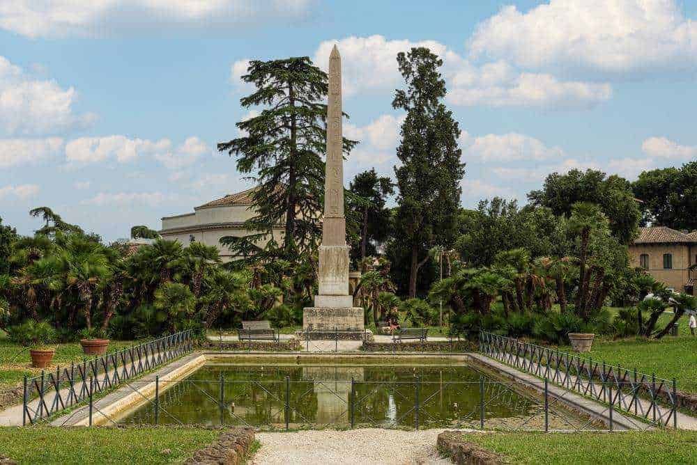 Antico Obelisco in una piazza storica con fontana, alberi maestosi e panchine, ambiente tranquillo e pittoresco, ideale per passeggiate e relax in una zona verde di Roma.