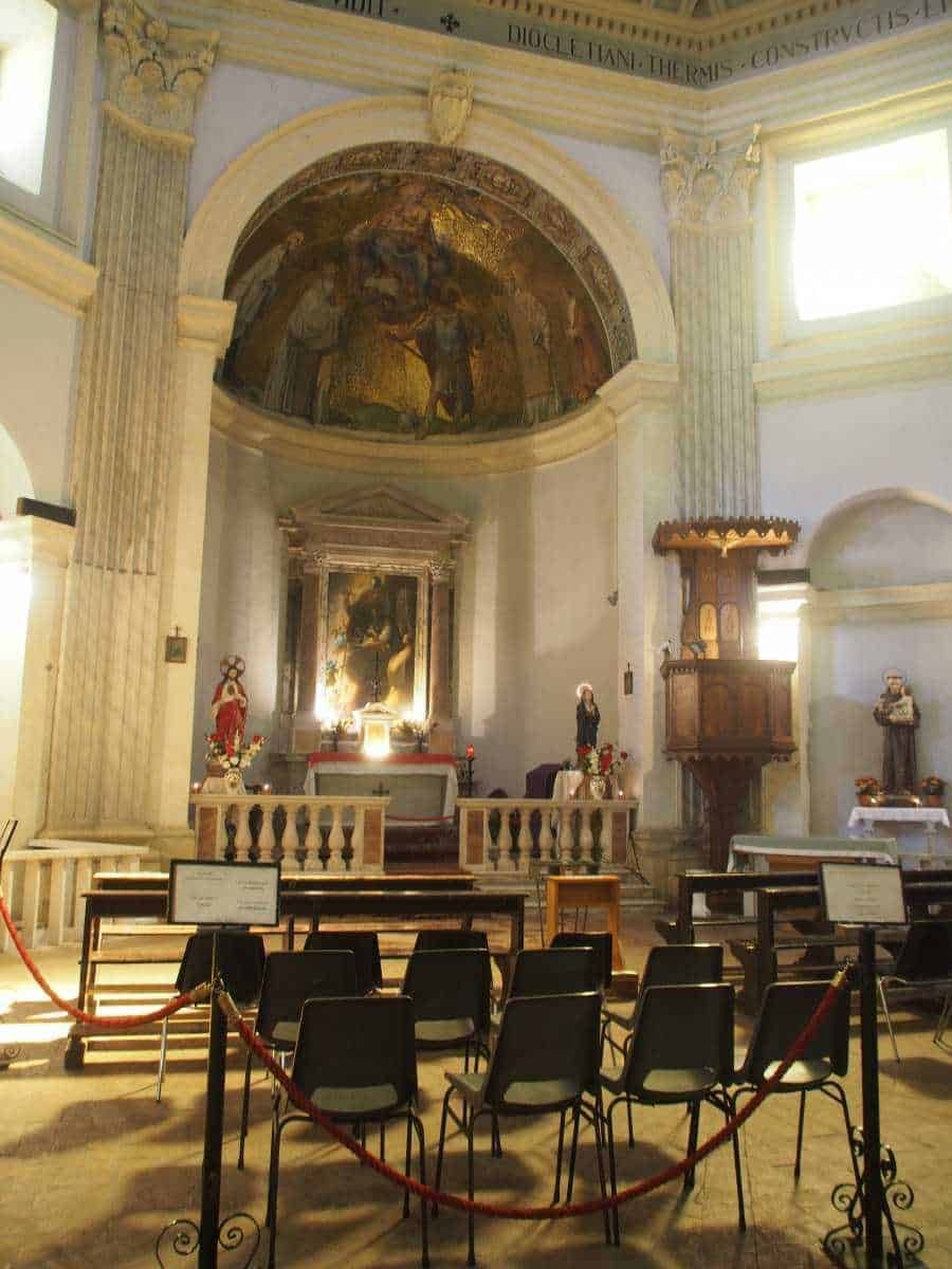 Antico luogo di culto con arredi sacri, affreschi storici e sedie in sala, decorato con statue religiose e cornici in legno, illuminato dalla luce naturale delle finestre.