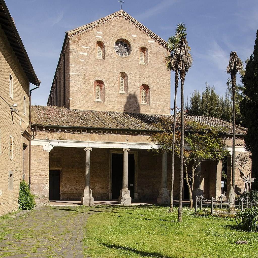 Scorcio della chiesa romanica con portico e colonne antiche, circondata da palme e alberi, in un cortile con calpestio in pietra e verde ben curato, cielo sereno.