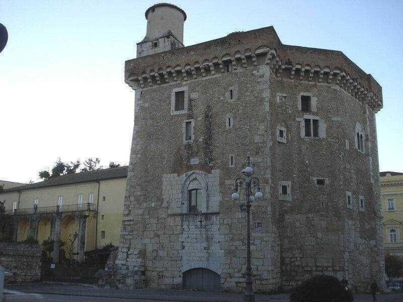 Antica torre medievale in pietra con torrette e finestre storiche, situata in un centro urbano italiano, simbolo di architettura storica e cultura locale.