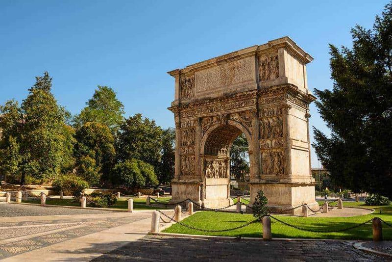 Arco di Tito a Roma situato in un parco verde con alberi e sentieri, sotto un cielo azzurro, rappresenta un antico monumento romano e un importante sito storico italiano.