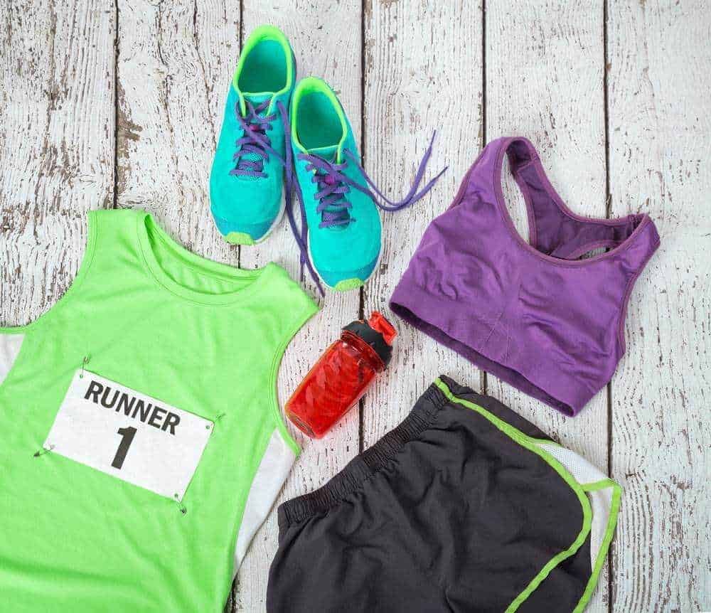Scarpe sportive da corsa, canotta da running, borraccia, pantaloncini e attrezzatura fitness su sfondo in legno bianco, perfetti per allenamenti all'aperto e attività sportive.