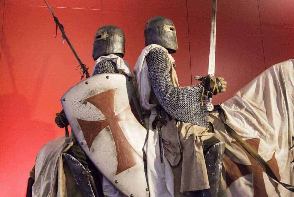 Armature da cavaliere templare con scudo crociato e spada, vestiti in abiti medievali, posizionata come statua o esposizione storica, sfondo rosso.