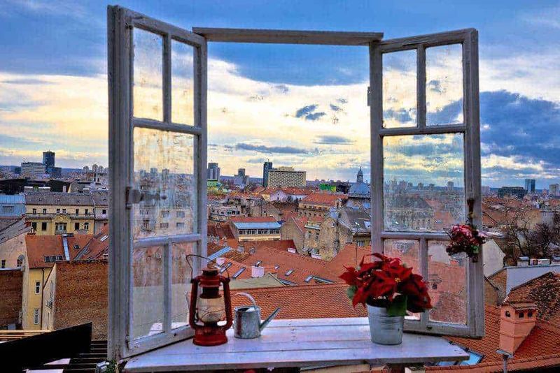 Vetra aperta su tetto di città con case in stile europeo e cielo nuvoloso, atmosfera pittoresca e suggestiva, panoramica urbana dall'antico finestrino di legno.