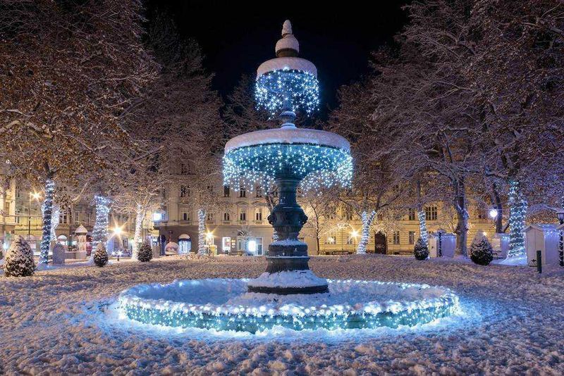 Fontana decorata con luci natalizie nella neve di un parco in notturna, con alberi e palazzi illuminati. Atmosfera festiva e magica, ideale per feste di Natale e decorazioni invernali.
