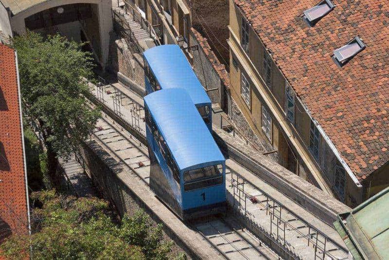 Luce di un tram blu storico in centro città che attraversa strade suggestive con edifici storici e tetti in tegole rosse. Scatto aereo che cattura la vita urbana e il trasporto pubblico vintage.