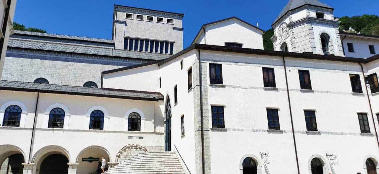 Antico monastero con archi e torre in pietra bianca, situato in un sito storico italiano, circondato da colline verdi e cielo blu, perfetto esempio di architettura religiosa e patrimonio culturale.