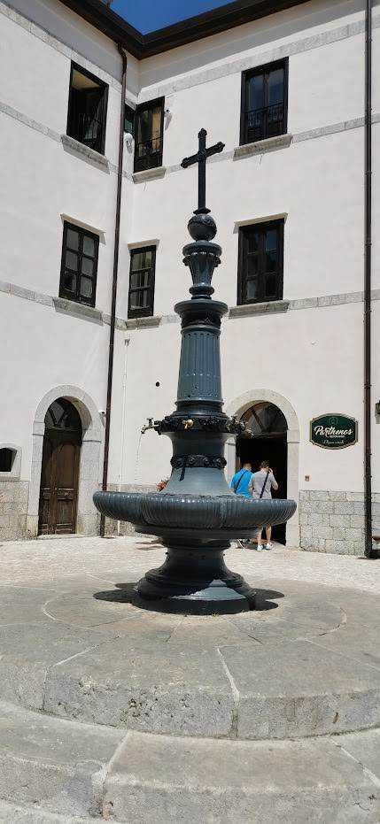 Fontana storica in piazza nel centro storico di una città italiana, con edificio bianco sullo sfondo, persone che passeggiano e atmosfera tradizionale e culturale.