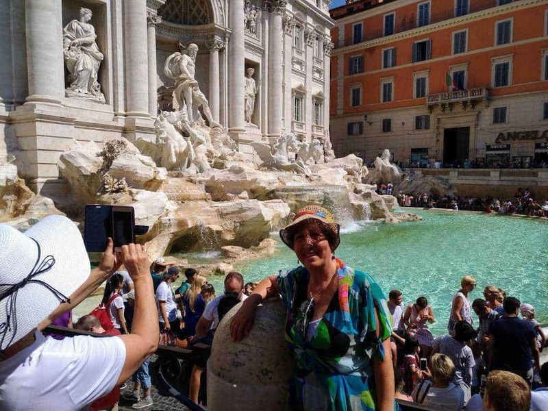 Perché è famosa la Fontana di Trevi? 2 Femminile turista sorridente davanti alla Fontana di Trevi a Roma, Italia, mentre scatta una foto, con molti visitatori e architetture storiche sullo sfondo.