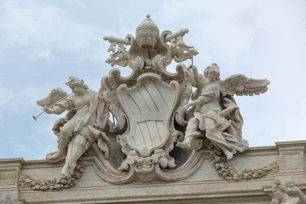 Fantasioso stemma araldico in scultura di marmo con due Angeli alati decorativi e dettagli artistici su un edificio storico, rappresentato come esempio di arte barocca italiana.