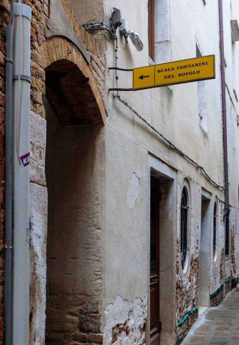Scala del Bovolo a Venezia, tra architettura gotica: dove si trova? 5 Vecchio vicolo medievale con segnale stradale giallo "Scala Contarini del Bovalo" e facciata in pietra e intonaco decorativo.