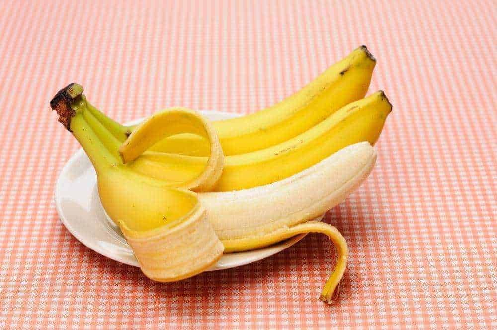 Banana con buccia parzialmente rimossa, su piatto bianco su tovaglietta a quadri color pesca, close-up.