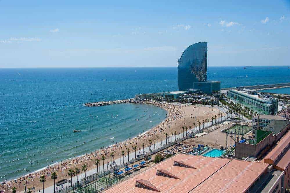 Spiaggia di Barcellona con La Sagrada Família sullo sfondo, vista dal mare con ombrelloni e persone che si rilassano sulla sabbia, mare cristallino e cielo sereno, destinazione turistica popolare per le vacanze estive.