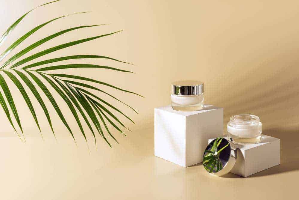 Creme viso skincare in vasi di vetro su supporti bianchi con foglia di palma verde sullo sfondo naturale e minimalista.