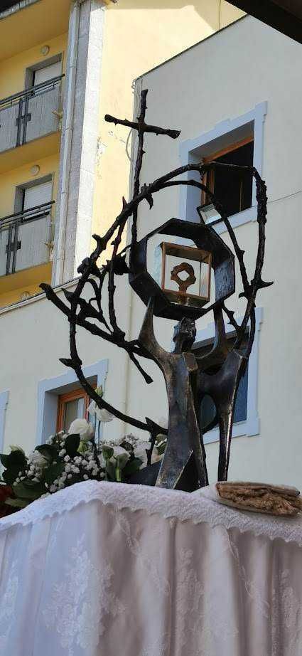 Scultura religiosa in metallo con croce e cuore, decorata con fiori bianchi, allestimento per celebrazione religiosa in spazio urbano.