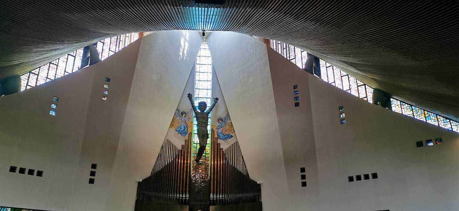 Finestra e interno di una chiesa moderna con scultura di Cristo sulla croce, pareti bianche e grandi vetrate colorate che permettono la luce naturale di entrare. Questo spazio sacro unisce arte contemporanea e architettura religiosa.