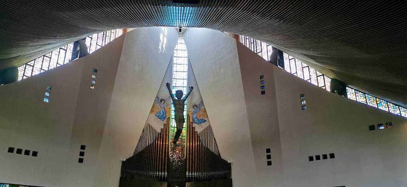 San Gerardo Maiella protettore dei bambini 8 Finestra e interno di una chiesa moderna con scultura di Cristo sulla croce, pareti bianche e grandi vetrate colorate che permettono la luce naturale di entrare. Questo spazio sacro unisce arte contemporanea e architettura religiosa.
