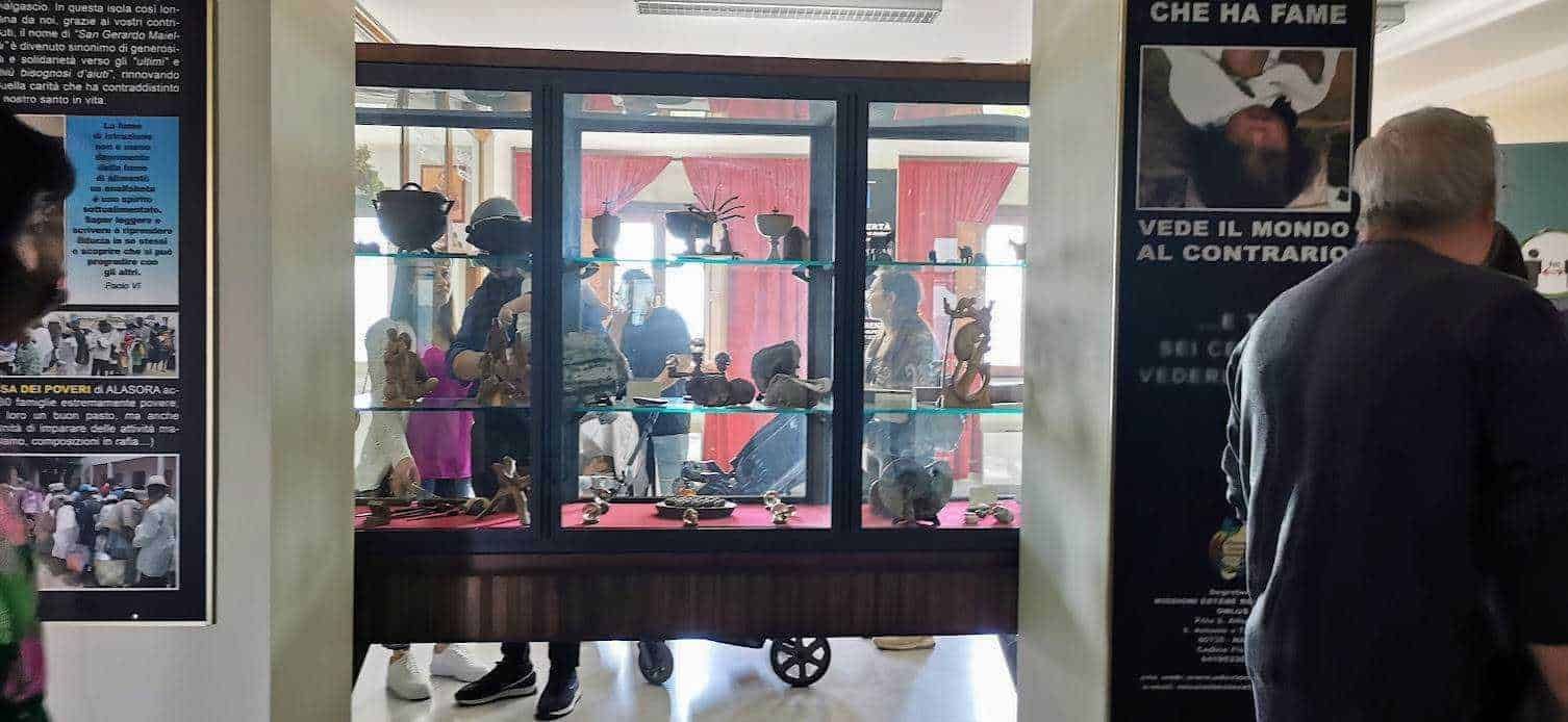 Vetrina di oggetti d'artigianato e sculture in esposizione all'interno di un negozio di antiquariato, visibile attraverso una grande finestra di vetro. La scena è animata da visitatori e clienti che osservano e acquistano pezzi unici.