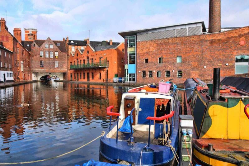 13 Città con canali nel mondo da vedere 5 Barche attraccate su un canale di una città storica con edifici in mattoni rossi e un ponte in pietra, scena urbana tipica di un quartiere antico ricco di architettura e cultura.