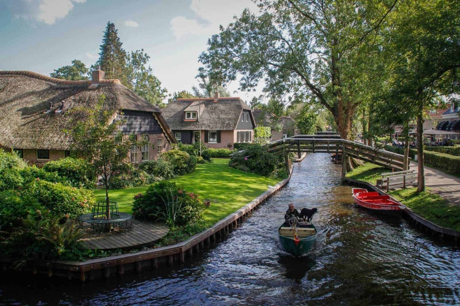 Casa olandese in stile tradizionale lungo un canale con giardino curato e barche in acqua, in un paesaggio pittoresco tipico delle città olandesi, con alberi verdi e cielo sereno.