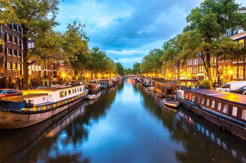 13 Città con canali nel mondo da vedere 3 Barche sul fiume durante il tramonto in un quartiere storico di Amsterdam, con case tradizionali e alberi lungo le rive, creando un'atmosfera suggestiva e pittoresca.
