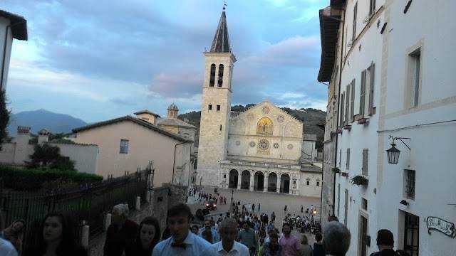 Campanile e facciata della chiesa di San Michele a Borgo San Michele, Italia, con molte persone e un cielo nuvoloso sullo sfondo.