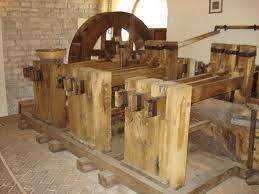 Antico mulino a vento e ingranaggi in legno, simbolo di tradizione e artigianato italiano, con un vecchio uomo che lavora davanti a un fiume in cascata, e un bambino che scrive su un quaderno a scuola.