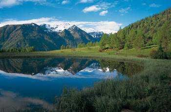 Lago alpino circondato da montagne verdeggianti e neve, riflesso limpido in acqua.