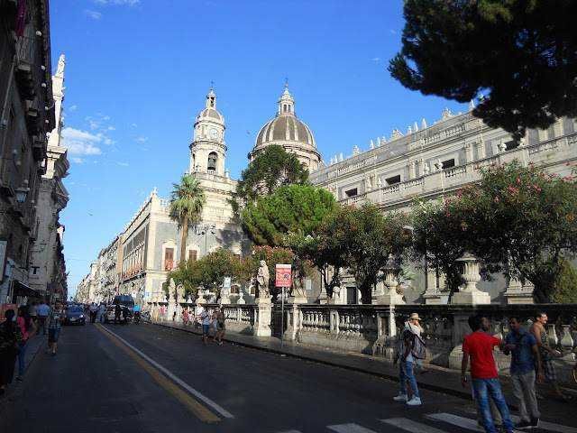 Catania, gita di un giorno percorrendo una sola strada Elegante vista di un edificio storico con cupole e architettura classica, circondato da alberi e persone che passeggiano in strada, in una giornata soleggiata e serena in centro città.