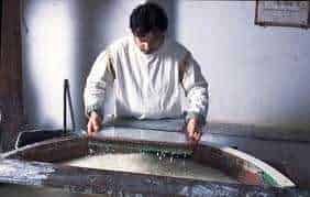 Antico mulino a vento e ingranaggi in legno, simbolo di tradizione e artigianato italiano, con un vecchio uomo che lavora davanti a un fiume in cascata, e un bambino che scrive su un quaderno a scuola.